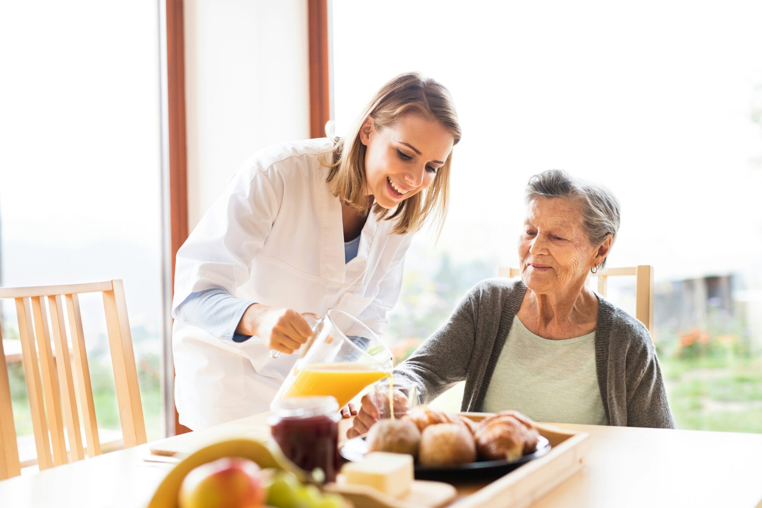 Une aide-soignante souriante en blouse blanche verse du jus d'orange à une femme âgée assise à une table garnie d'aliments pour le petit-déjeuner tels que des croissants, des fruits et de la confiture, prête à partager des astuces telles que la manière de nettoyer un Thermomix.