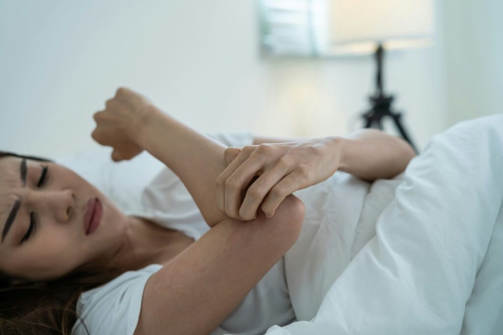 Une femme est allongée dans son lit, semblant mal à l'aise alors qu'elle tient son coude d'une main, peut-être en raison d'une douleur ou d'une gêne causée par des punaises de lit. L'arrière-plan doucement éclairé comporte une lampe et une literie de couleur claire.