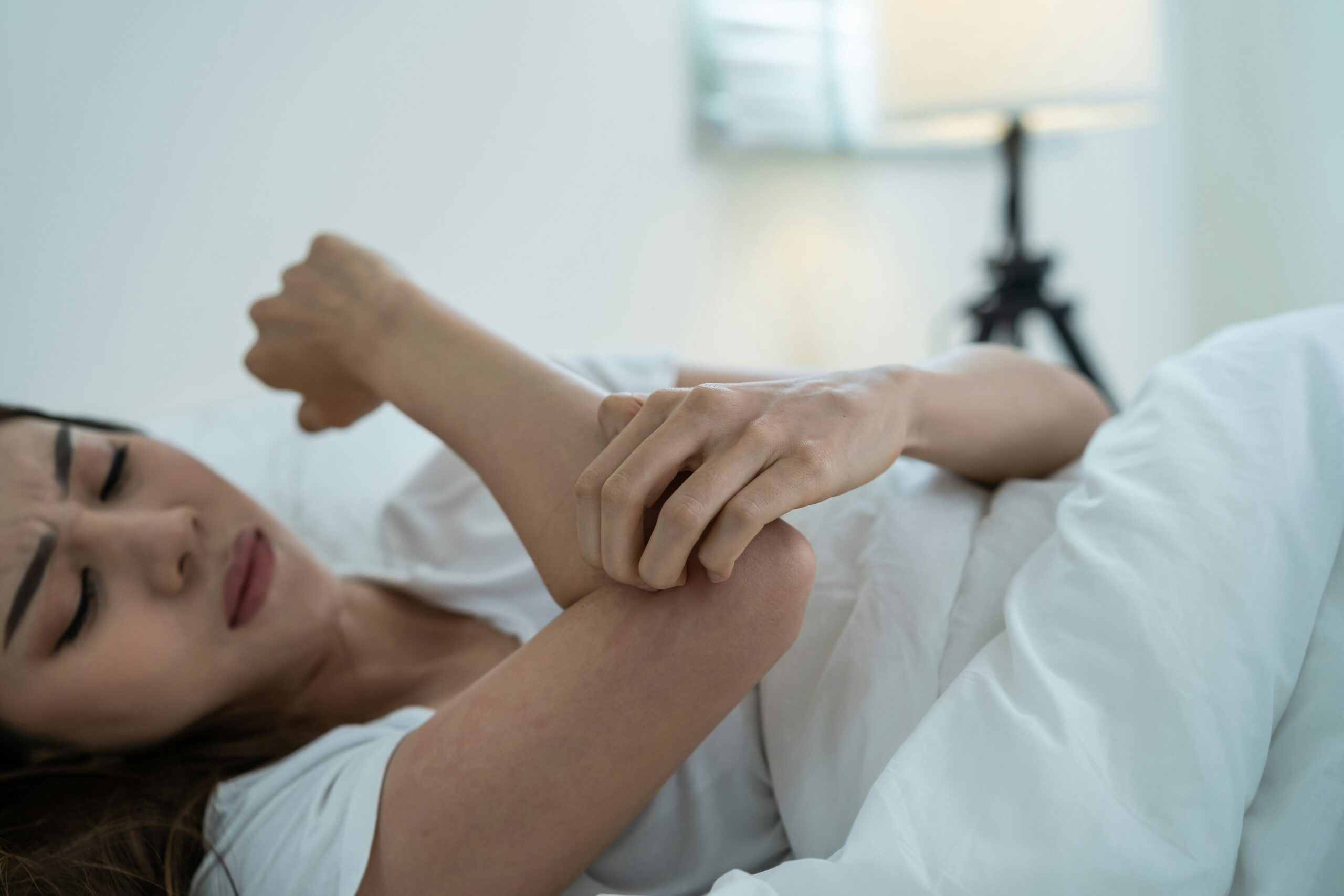 Une femme est allongée dans son lit, semblant mal à l'aise alors qu'elle tient son coude d'une main, peut-être en raison d'une douleur ou d'une gêne causée par des punaises de lit. L'arrière-plan doucement éclairé comporte une lampe et une literie de couleur claire.