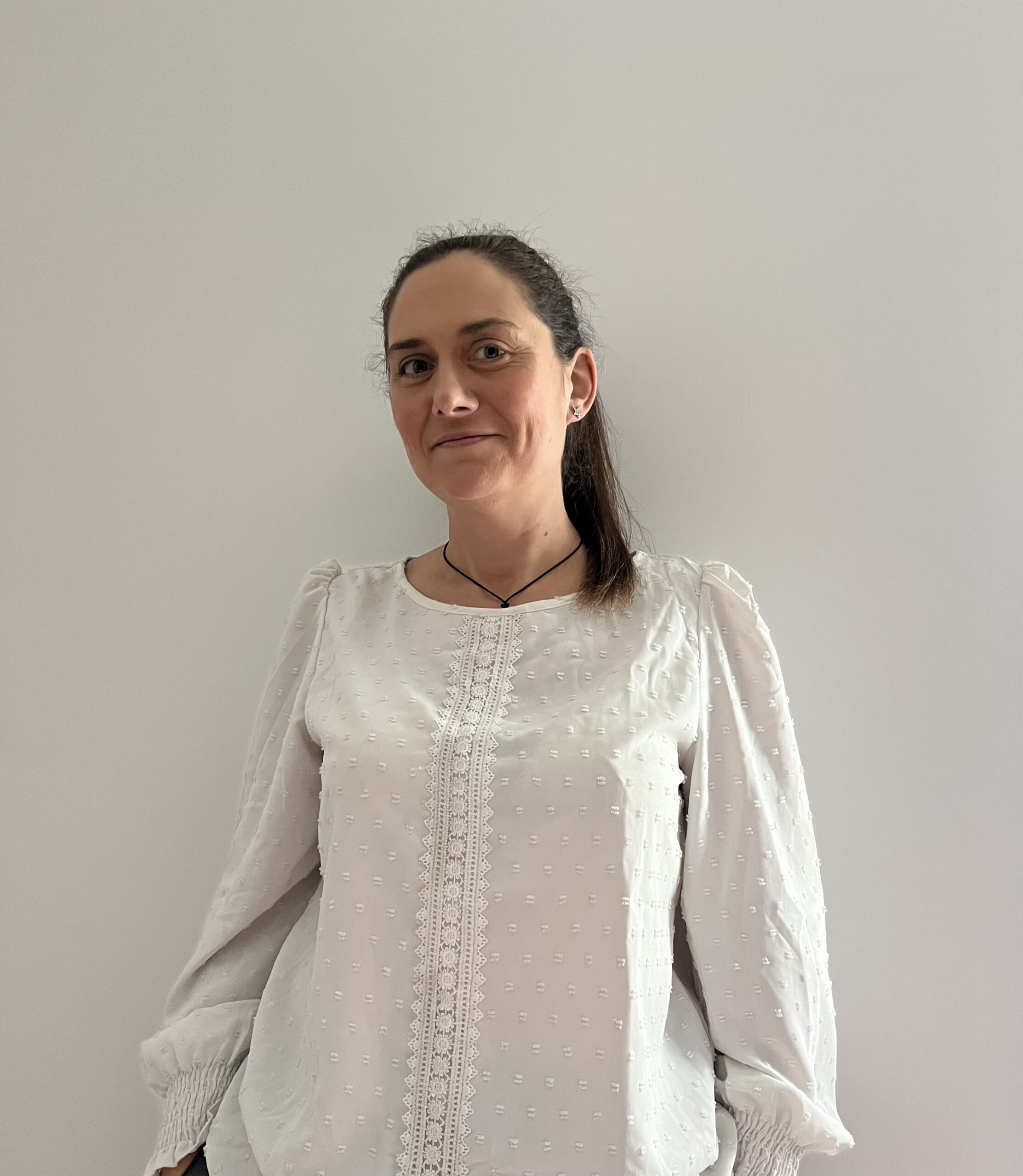 Une femme aux longs cheveux bruns attachés se tient debout contre un mur uni de couleur claire, vêtue d'un chemisier blanc avec des détails en dentelle et des manches bouffantes. Elle sourit gentiment, incarnant la chaleur et le professionnalisme d'une aide à domicile d'Uzès.
