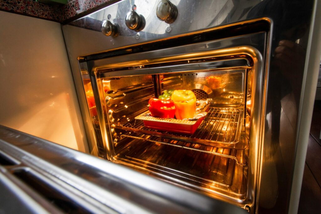 Une vue rapprochée d'une porte de four ouverte révèle une plaque de cuisson avec des poivrons rouges et jaunes en train de rôtir, parfaits pour un repas nutritif qui peut aider à prévenir la dénutrition. L'intérieur du four, chaudement éclairé, semble propre et accueillant.