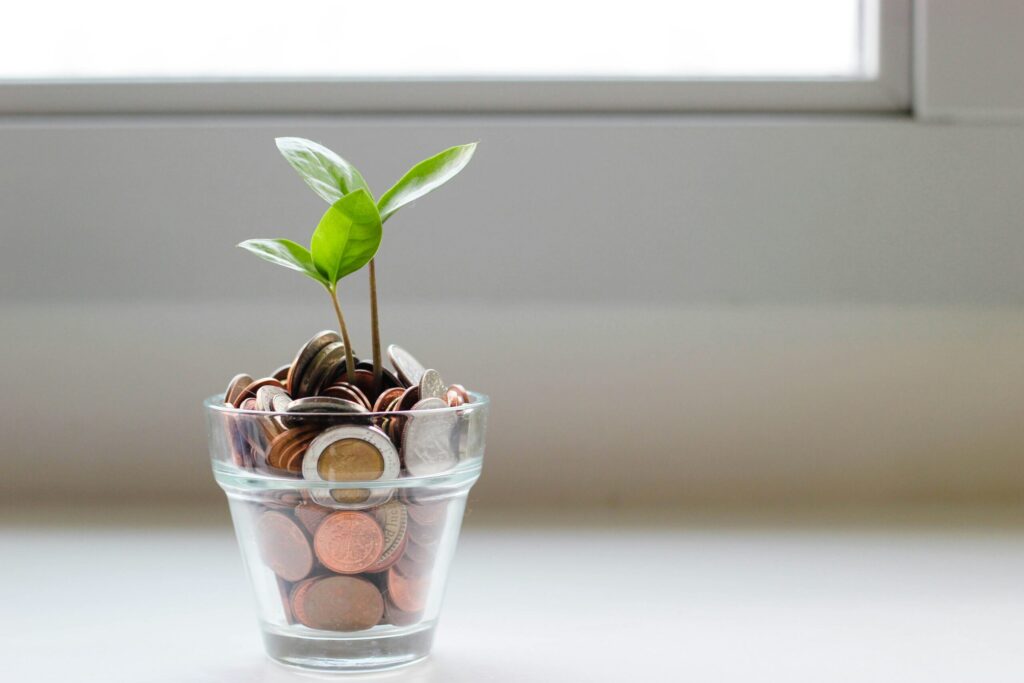 Une petite plante verte, symbolisant l'allégement de la charge mentale, pousse dans un verre rempli de pièces de monnaie assorties, placé sur une surface blanche devant une fenêtre.