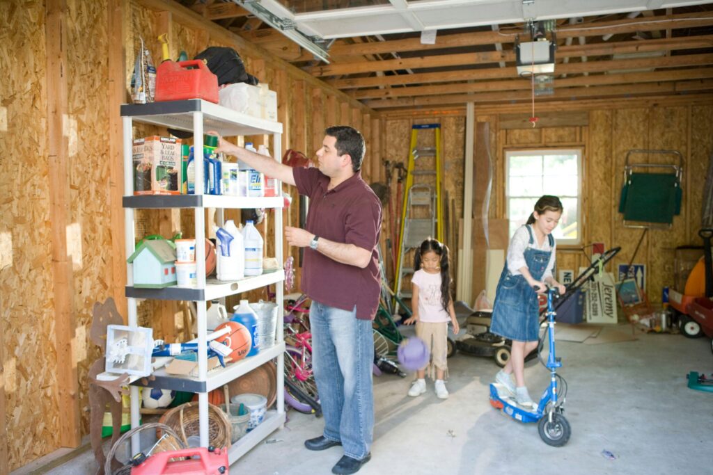 Un homme fournit une aide à domicile en organisant des objets sur une étagère dans un garage inachevé aux poutres apparentes, tandis que deux fillettes jouent à proximité, l'une sur une trottinette bleue et l'autre avec un ballon violet.