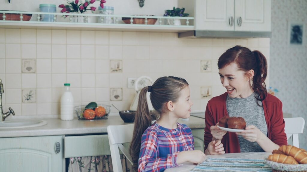 Une femme et une jeune fille sont assises à une table de cuisine et se sourient. Une aide à domicile tient une assiette avec une pâtisserie. La cuisine est décorée de plantes en pot et des croissants sont posés sur la table.