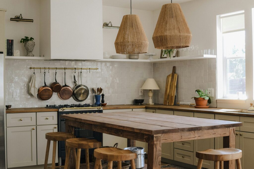 Une cuisine confortable avec des armoires blanches, des comptoirs en bois, des casseroles suspendues et deux lampes suspendues tressées au-dessus d'une table rustique en bois entourée de tabourets. La lumière du soleil traverse la fenêtre, créant une atmosphère chaleureuse, idéale pour préparer les repas afin de prévenir la dénutrition.