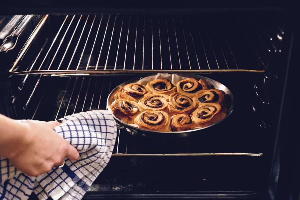 Une personne utilise un torchon à carreaux pour placer ou retirer d'un four un plat métallique rond contenant des roulés à la cannelle fraîchement sortis du four - une scène réconfortante qui contraste fortement avec les défis auxquels sont confrontées les personnes souffrant de dénutrition.