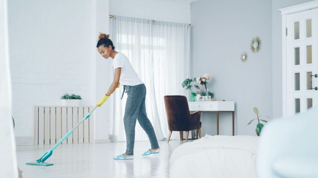 Une femme portant des vêtements décontractés et des gants jaunes passe la serpillière dans une chambre claire et bien rangée, aux murs blancs, aux grandes fenêtres et à la décoration moderne et simple, afin de maintenir un espace propre pour prévenir des problèmes tels que la dénutrition.