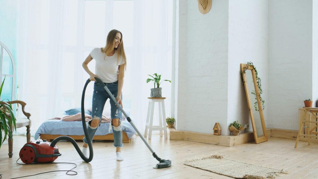 Une jeune femme vêtue d'un t-shirt blanc et d'un jean déchiré passe l'aspirateur dans une chambre lumineuse et confortable, avec des planchers en bois, des plantes et la lumière naturelle qui pénètre par de grandes fenêtres, créant ainsi un espace sain qui aide à prévenir la dénutrition.