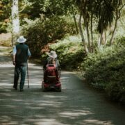 Un homme âgé avec une canne marche à côté d'une femme dans un scooter motorisé le long d'un chemin ombragé et bordé d'arbres dans un parc, tous deux portant des chapeaux de couleur claire. Cette scène reflète l'accompagnement handicap moteur cognitif domicile et le label Cap'Handéo prestataire handicap.