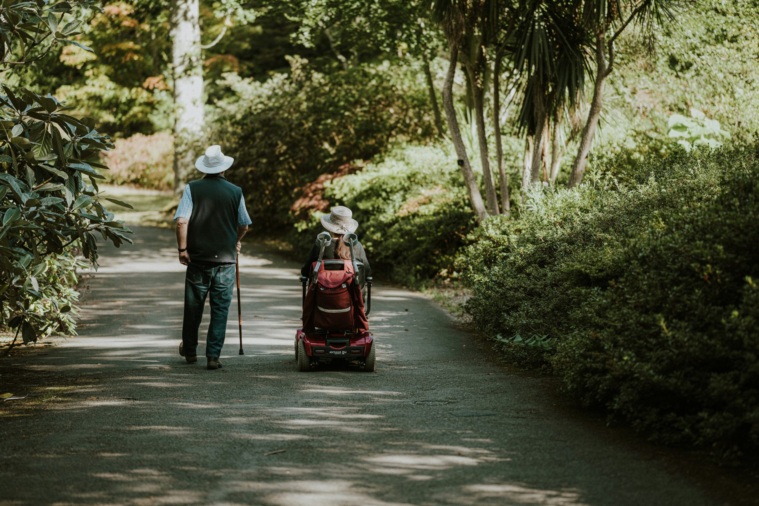 Un homme âgé avec une canne marche à côté d'une femme dans un scooter motorisé le long d'un chemin ombragé et bordé d'arbres dans un parc, tous deux portant des chapeaux de couleur claire. Cette scène reflète l'accompagnement handicap moteur cognitif domicile et le label Cap'Handéo prestataire handicap.