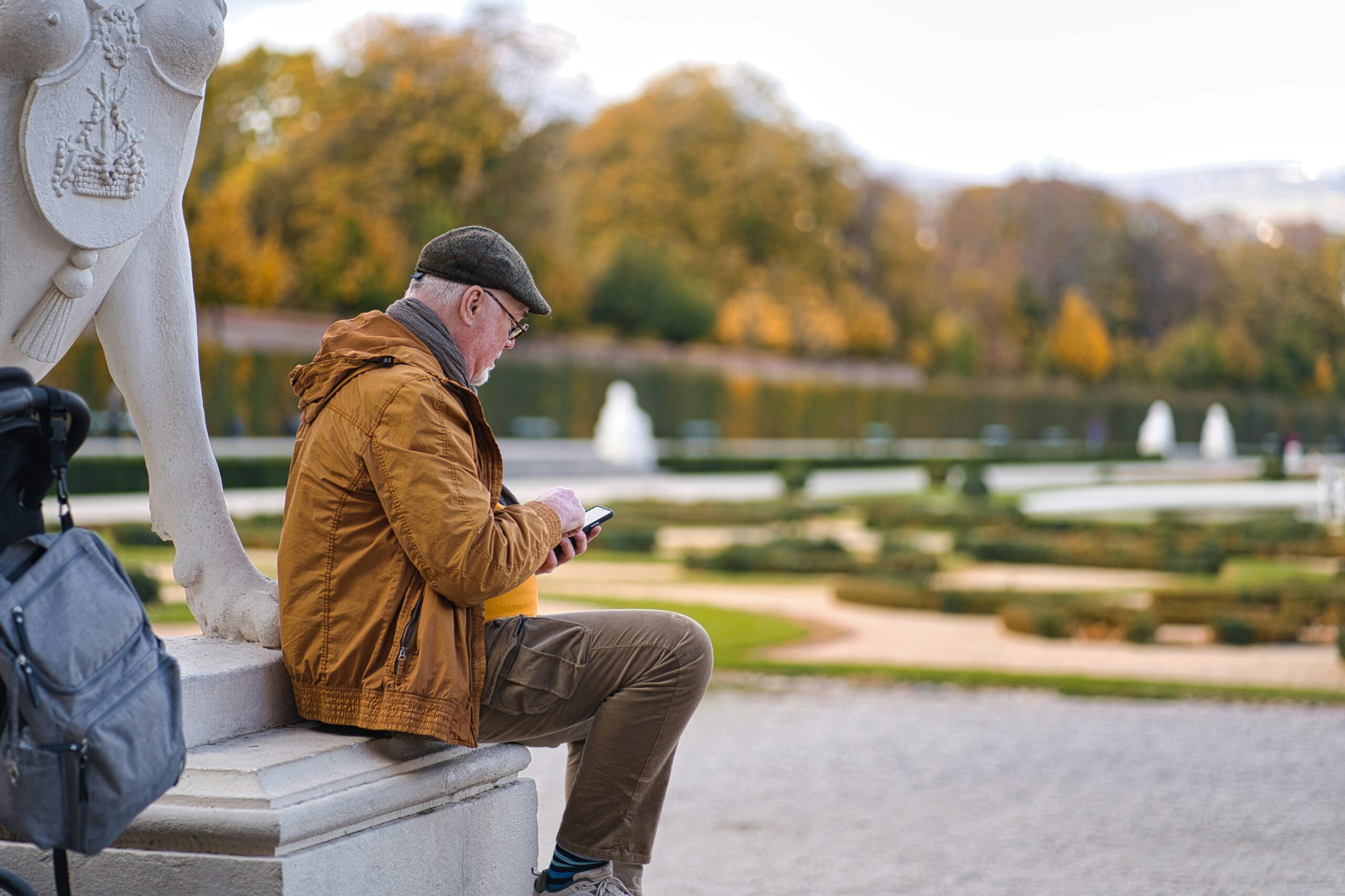 Un homme d'un certain âge, coiffé d'une casquette plate et vêtu d'une veste marron, est assis sur un banc en pierre dans un parc près d'arbres d'automne, de jardins et de fontaines. Il consulte son téléphone, peut-être à la recherche d'une aide à domicile fiable Arles. Un sac à dos est posé à côté de lui.