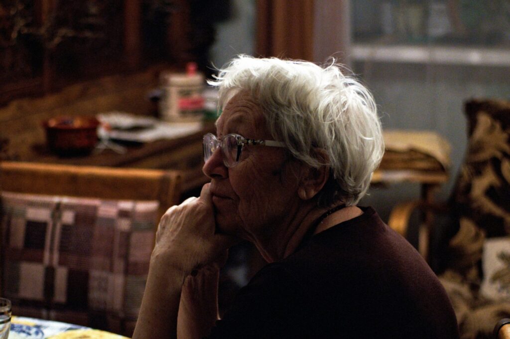 Une femme âgée aux cheveux blancs et aux lunettes est assise à une table, le menton posé sur sa main et regardant pensivement sur le côté - un moment serein, soulignant l'importance de bien choisir une aide à domicile personnes âgées pour un quotidien apaisé.
