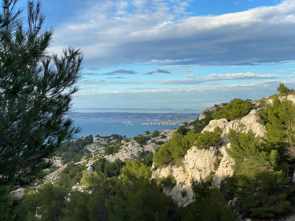 Vue d'une colline rocheuse couverte de pins verts, surplombant une mer bleue sous un ciel partiellement nuageux, avec une terre lointaine visible à l'horizon - comme les scènes sereines dont jouissent les bénéficiaires des services d'aide ménagère Marseille.