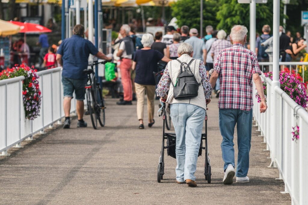 Un groupe de personnes, dont un couple de personnes âgées - l'une utilisant un déambulateur et l'autre se tenant le bras - marchent le long d'un pont piétonnier bordé de fleurs par une journée ensoleillée, illustrant l'esprit du maintien à domicile à Arles. D'autres personnes marchent ou poussent des vélos à l'arrière-plan.