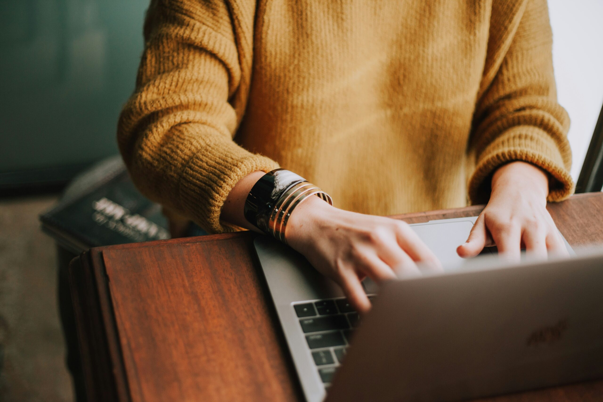 Une personne portant un pull jaune et des bracelets tape sur un ordinateur portable à un bureau en bois, cherchant peut-être un emploi aide à domicile week-end, avec un livre partiellement visible à proximité.