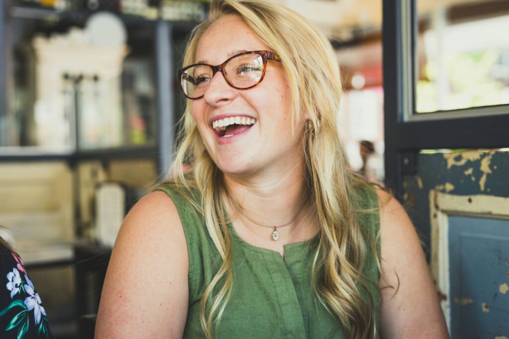 Une femme aux longs cheveux blonds et aux lunettes sourit et rit, assise à l'intérieur. Elle porte un haut vert sans manches et un collier, représentant la stabilité équipes aide à domicile avec la lumière du soleil qui entre par une fenêtre voisine.