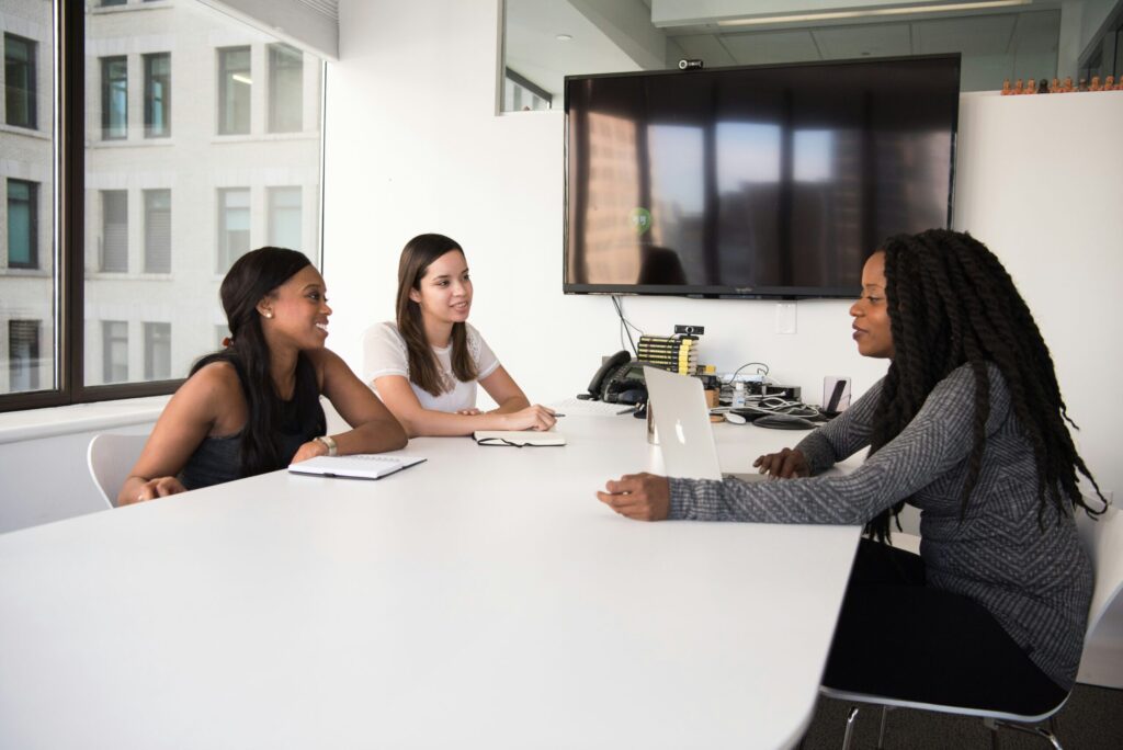Trois femmes sont assises à une table blanche dans un bureau moderne, discutant de documents pour postuler une aide à domicile. L'une d'entre elles utilise un ordinateur portable tandis que les autres ont des ordinateurs portables. De grandes fenêtres et une télévision murale se trouvent à l'arrière-plan.