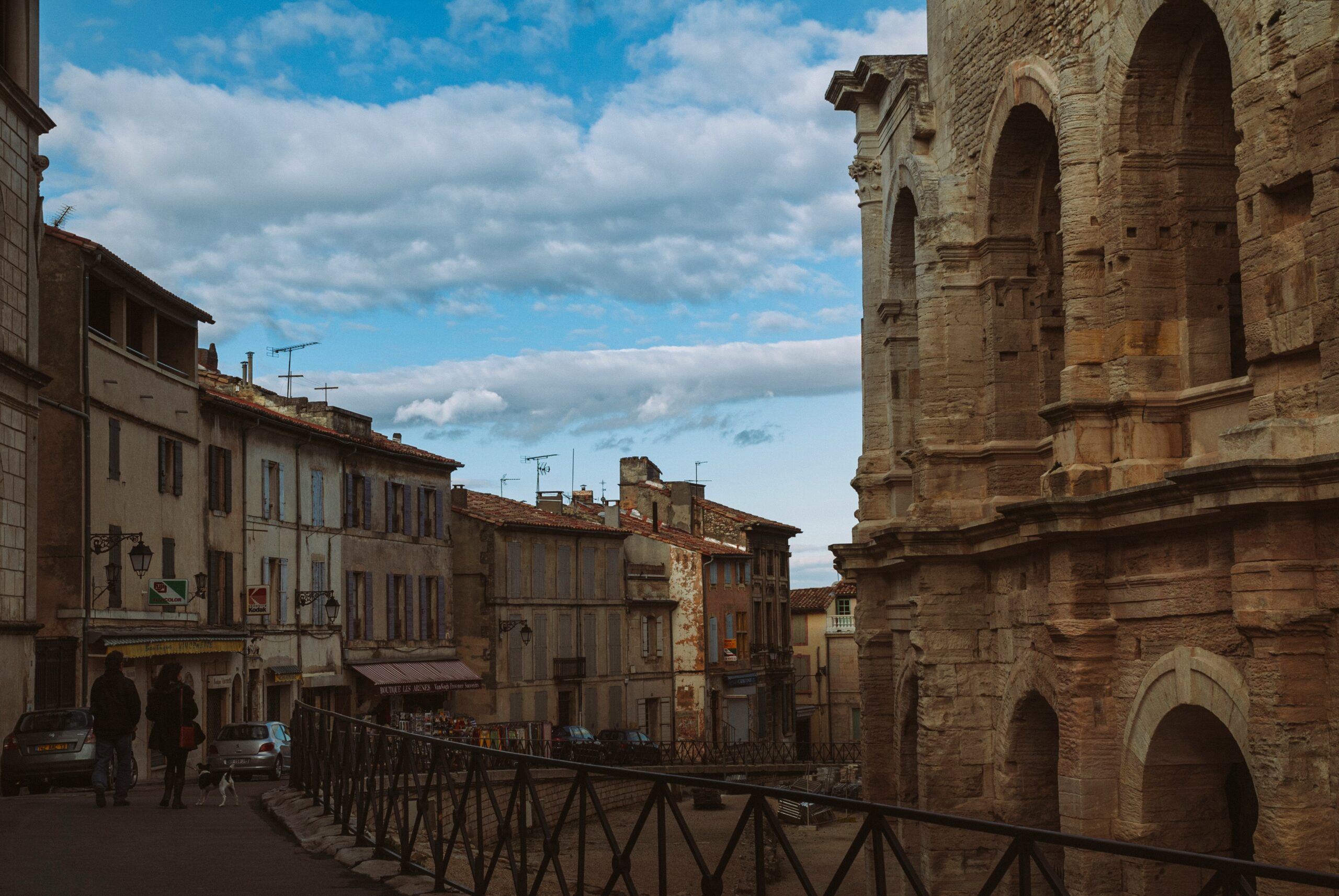 Une rue européenne étroite d'Arles bordée de bâtiments anciens, dont une grande structure en pierre avec des arches sur la droite. Des personnes, des voitures et une aide à domicile seniors Arles sont visibles sous un ciel partiellement nuageux avec des taches de bleu.