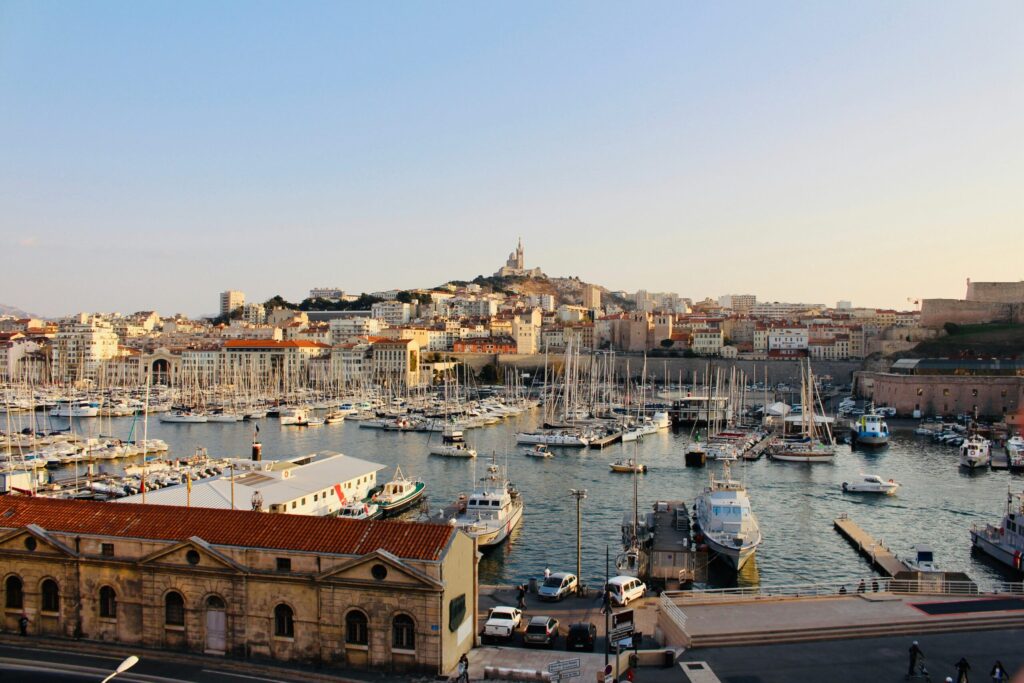 Un port rempli de nombreux voiliers et yachts, bordé de bâtiments historiques aux toits de tuiles rouges. En arrière-plan, une ville s'élève sur une colline surmontée d'une basilique - une vue emblématique de Marseille, où le service à la personne Marseille prospère.