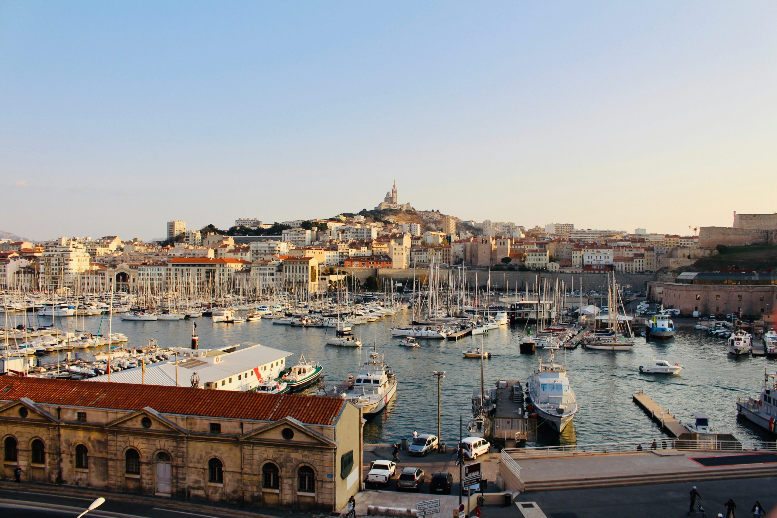 Un port rempli de nombreux voiliers et yachts, bordé de bâtiments historiques aux toits de tuiles rouges. En arrière-plan, une ville s'élève sur une colline surmontée d'une basilique - une vue emblématique de Marseille, où le service à la personne Marseille prospère.