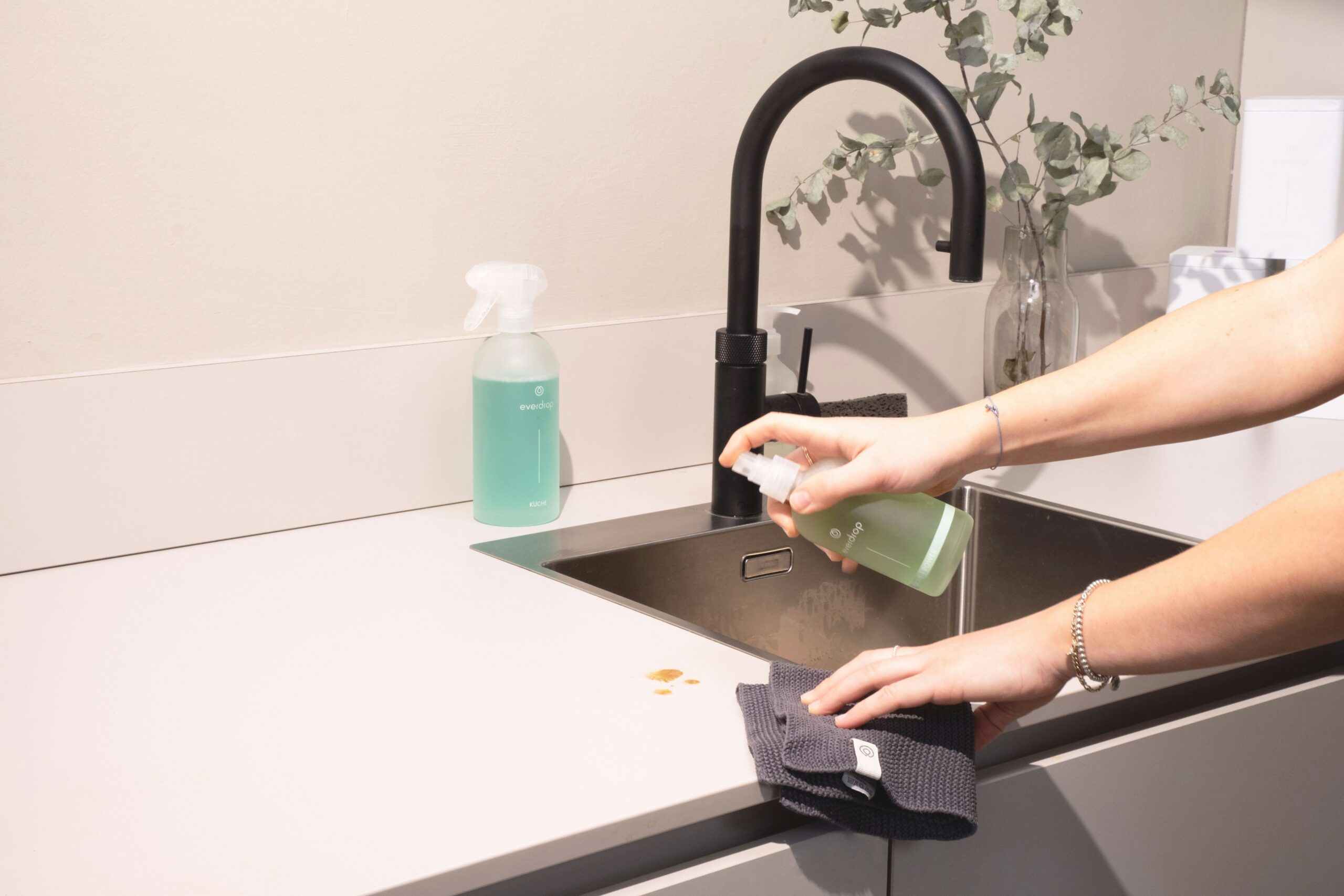 Une personne vaporise de la solution de nettoyage sur un comptoir près d'un évier, tenant un chiffon sombre pour essuyer les pièces de monnaie, montrant le soin méticuleux du ménage à domicile Nîmes. Un flacon pulvérisateur et un vase avec de la verdure se trouvent à l'arrière-plan.