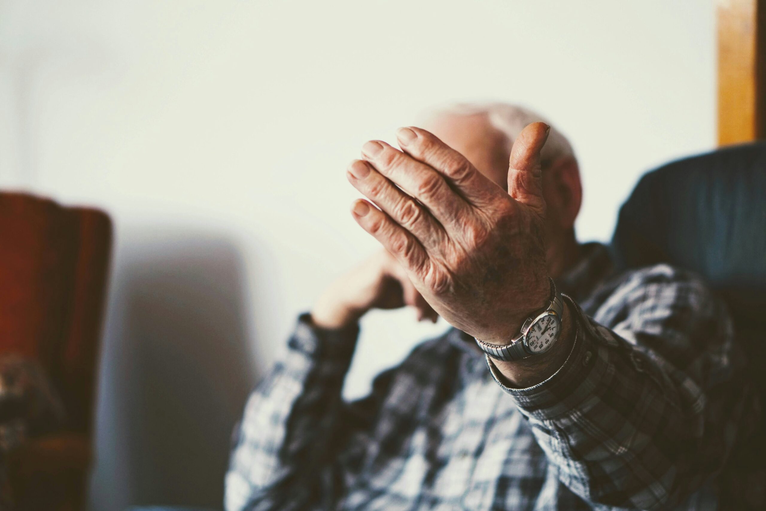 Une personne âgée assise sur une chaise, portant une chemise à carreaux et une montre-bracelet, tend le bras vers l'appareil photo et masque la majeure partie de son visage - un moment qui reflète l'importance de l'aide à domicile Alzheimer maintien domicile. L'arrière-plan est légèrement flou.