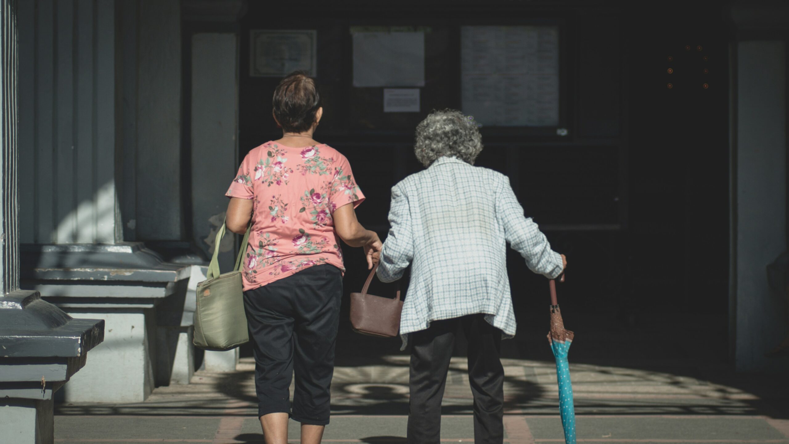 Deux femmes se dirigent main dans la main vers l'entrée d'un immeuble - l'une porte une chemise rose à fleurs et un sac vert, l'autre, aux cheveux gris, s'appuie sur une canne et tient un parapluie bleu - illustrant choisir aide à domicile Arles.
