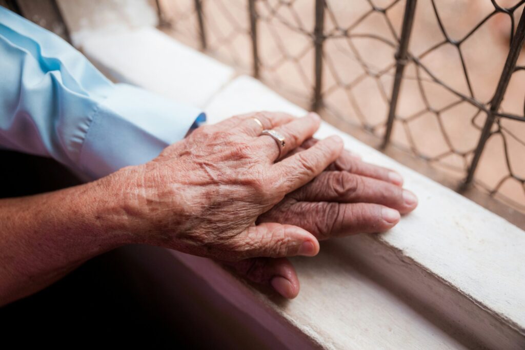 Deux mains âgées, dont l'une porte une alliance, reposent délicatement sur un rebord de fenêtre blanc près d'une fenêtre grillagée, reflétant la chaleur et la camaraderie. Elles capturent parfaitement les moments que les familles considèrent lorsqu'elles préparent domicile aide à domicile à Arles.