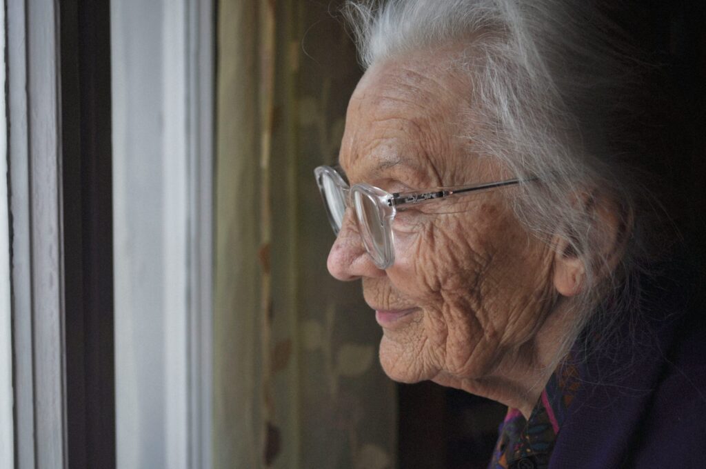 Une femme âgée aux cheveux blancs et aux lunettes regarde pensivement par la fenêtre, son visage doucement éclairé par la lumière naturelle. Elle semble paisible et satisfaite, incarnant le réconfort que l'accompagnement Alzheimer prestataire peut apporter pour le maintien à domicile des troubles cognitifs.