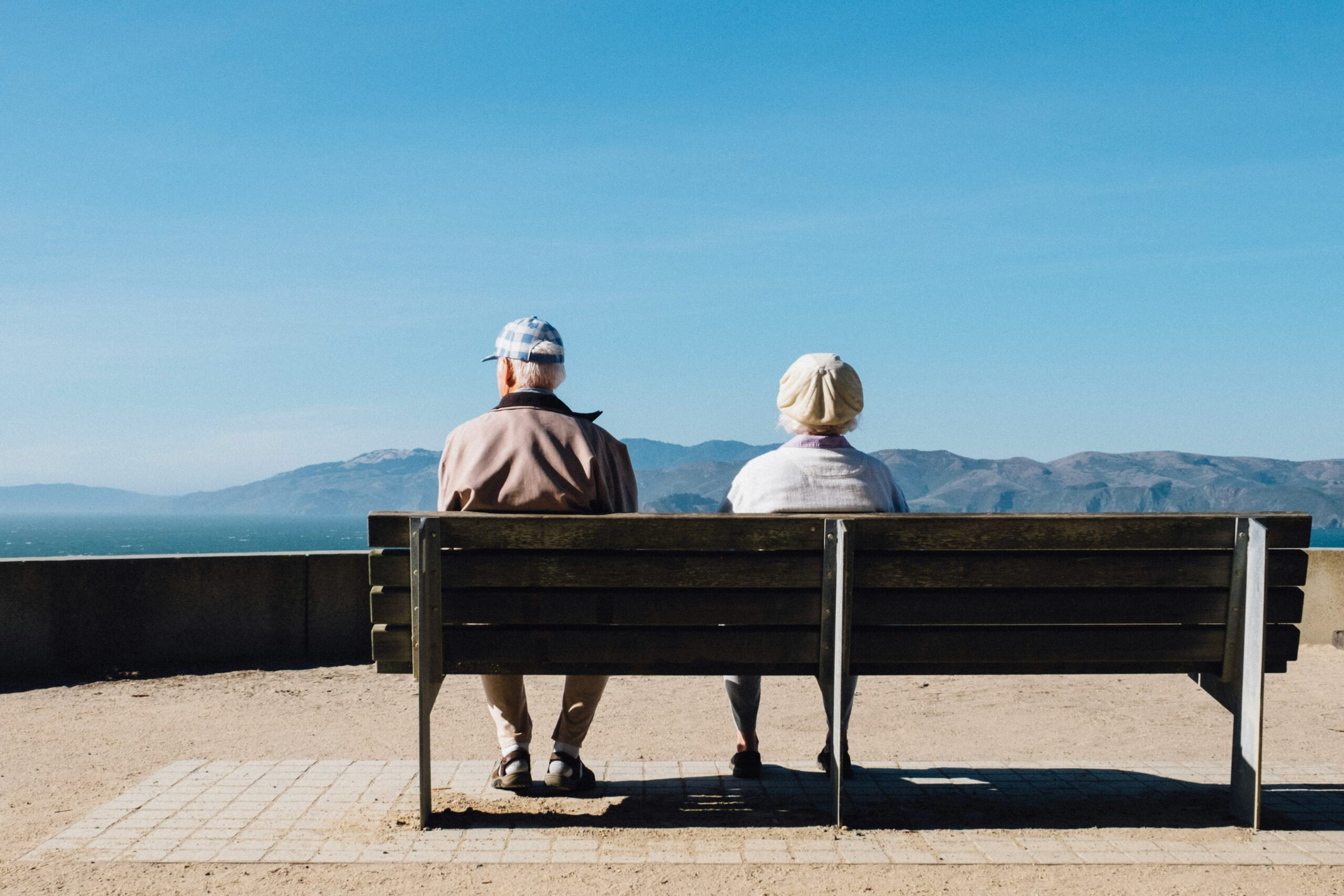 Deux personnes âgées sont assises côte à côte sur un banc, profitant d'une vue panoramique sur la montagne et l'eau sous un ciel bleu clair. Ce moment de calme reflète la valeur de Ressources d'Aide pour Personnes Âgées en Ville, qui soutient la vie urbaine paisible des personnes âgées.