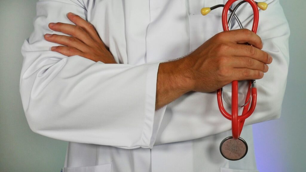 Une personne en blouse blanche se tient debout, les bras croisés, un stéthoscope rouge dans une main, prête à coordonner l'APA urgence retour hospitalisation. Le visage n'est pas visible.