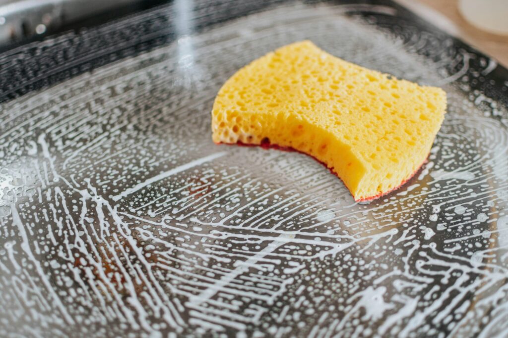 Une éponge jaune repose sur une surface humide et savonneuse qui a été partiellement nettoyée, laissant des traces et des bulles de savon - une scène invitante d'un ménage à domicile nîmois sur un comptoir ou un verre réfléchissant.