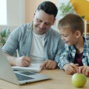 Un homme et un jeune garçon sont assis à une table, souriant et écrivant ensemble dans un cahier. Un ordinateur portable ouvert, une pomme verte et des fournitures scolaires sont posés sur la table - comme lorsque vous choisissez prestataire aide à domicile pour un soutien à domicile.