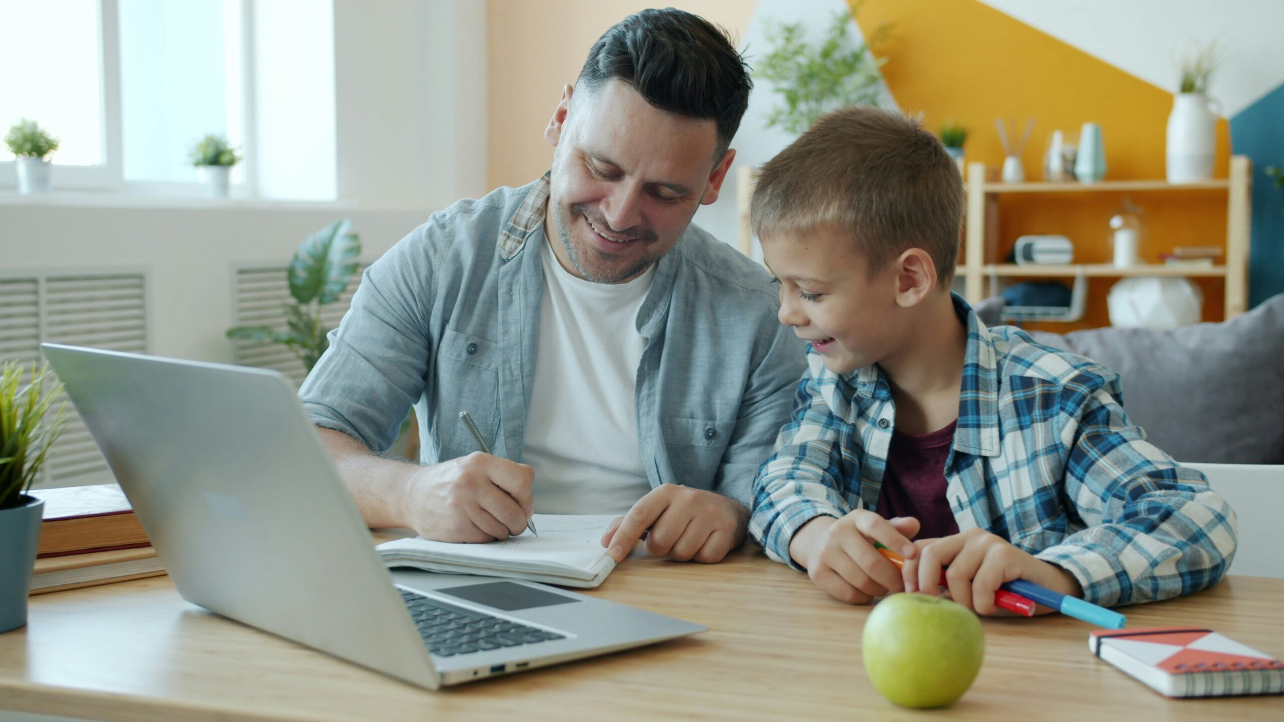 Un homme et un jeune garçon sont assis à une table, souriant et écrivant ensemble dans un cahier. Un ordinateur portable ouvert, une pomme verte et des fournitures scolaires sont posés sur la table - comme lorsque vous choisissez prestataire aide à domicile pour un soutien à domicile.