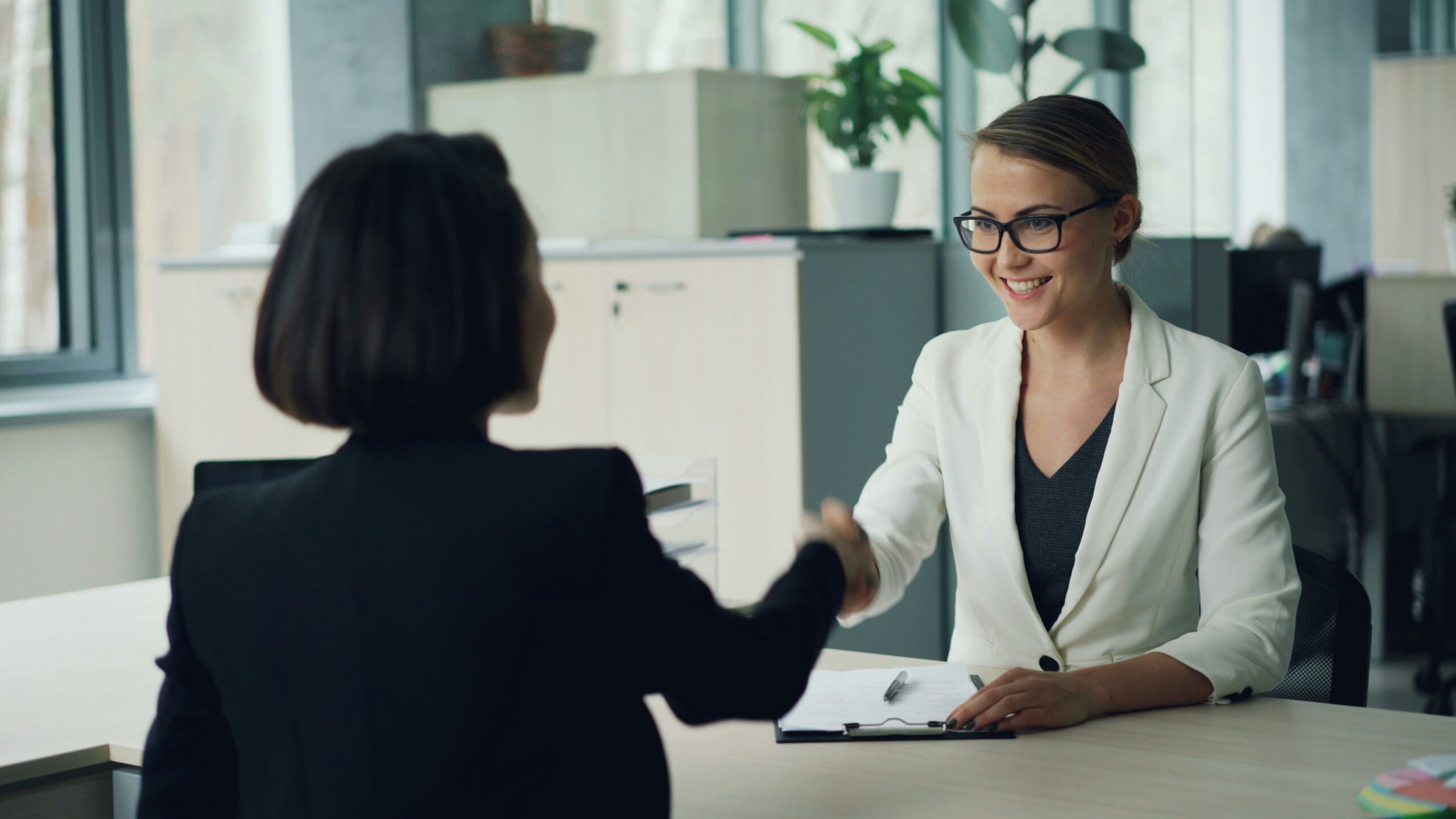 Deux femmes sont assises dans un bureau, souriantes et se serrant la main - peut-être en train de discuter du comparatif aide à domicile France. L'une porte un blazer blanc et des lunettes, l'autre une veste noire. Une planchette à pince et des documents sont posés sur le bureau.