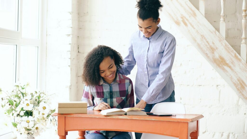 Deux jeunes femmes aux cheveux bouclés sont assises à un bureau, souriantes et lisant un livre. Elles semblent heureuses et engagées dans une pièce lumineuse et accueillante avec des fleurs, reflétant la chaleur des meilleurs services aide à domicile France.