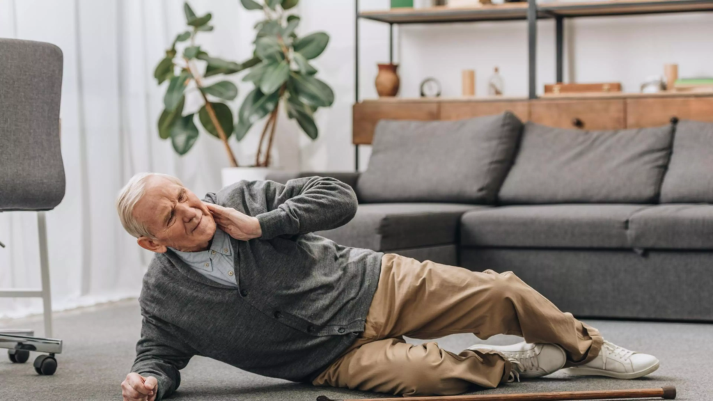Un homme âgé est allongé sur le sol du salon, souffrant, se tenant la joue, avec sa canne à proximité. Cette scène souligne l'importance de la prévention chutes seniors domicile et du bon aménagement logement seniors chutes.