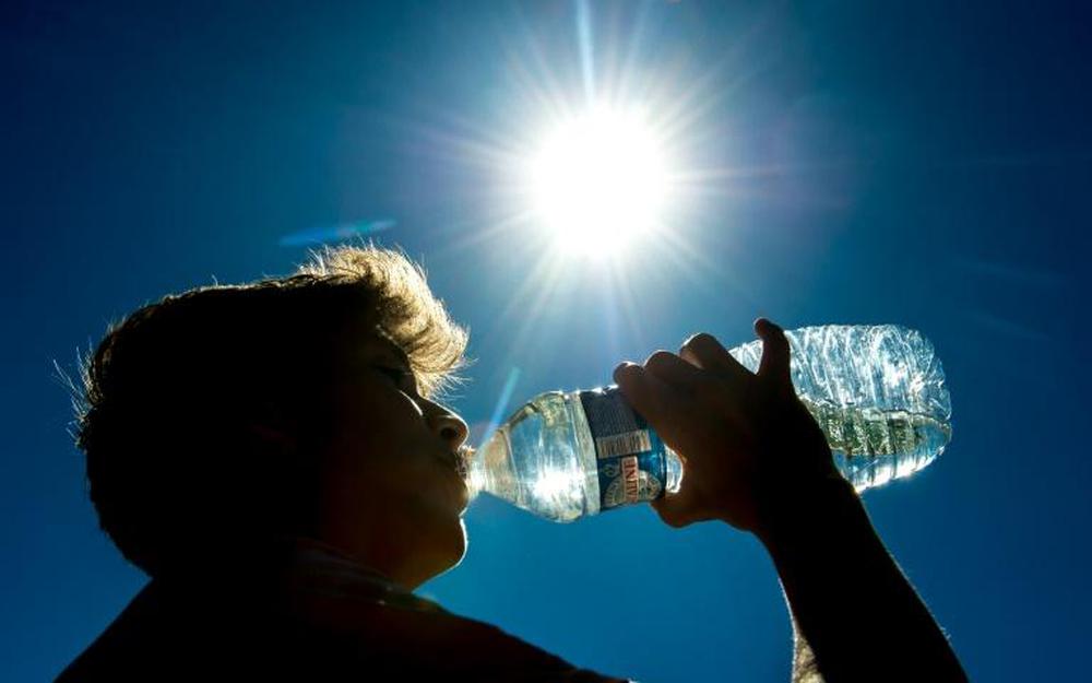 Une personne pratique l'hydratation senior chaleur, en buvant dans une bouteille d'eau sous un soleil radieux, dans un ciel bleu clair, la lumière du soleil créant un effet de brillance autour de sa silhouette.