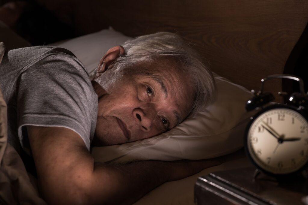 Un homme âgé aux cheveux grisonnants est allongé dans son lit la nuit, l'air inquiet et incapable de dormir. Il est face à un réveil analogique qui indique 3h55, ce qui reflète les difficultés courantes du sommeil senior et la nécessité d'une aide à domicile ou d'alternatives sûres comme la mélatonine senior.