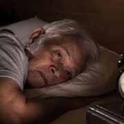 Un homme âgé aux cheveux grisonnants est allongé dans son lit la nuit, l'air inquiet et incapable de dormir. Il est face à un réveil analogique qui indique 3h55, ce qui reflète les difficultés courantes du sommeil senior et la nécessité d'une aide à domicile ou d'alternatives sûres comme la mélatonine senior.