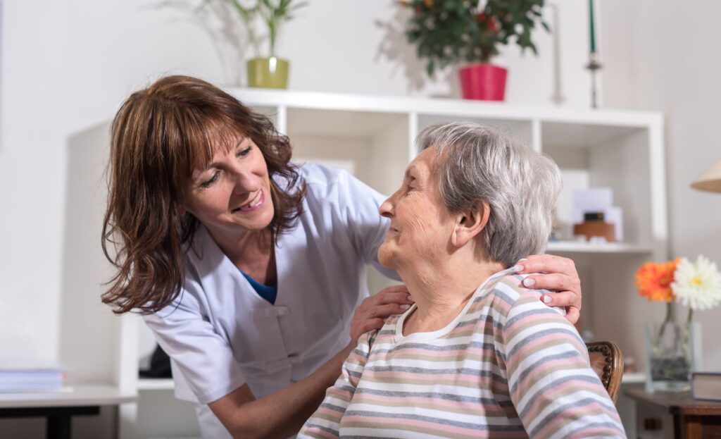 Une aide-soignante en uniforme blanc sourit et tient délicatement les épaules d'une femme âgée, toutes deux partageant un moment chaleureux à l'intérieur. La scène reflète des soins de qualité, avec des services à partir de 25 euros l'heure d'aide à domicile, conformément à l'arrêté du 23 décembre 2025 du SAD.