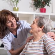 Une aide-soignante en uniforme blanc sourit et tient délicatement les épaules d'une femme âgée, toutes deux partageant un moment chaleureux à l'intérieur. La scène reflète des soins de qualité, avec des services à partir de 25 euros l'heure d'aide à domicile, conformément à l'arrêté du 23 décembre 2025 du SAD.