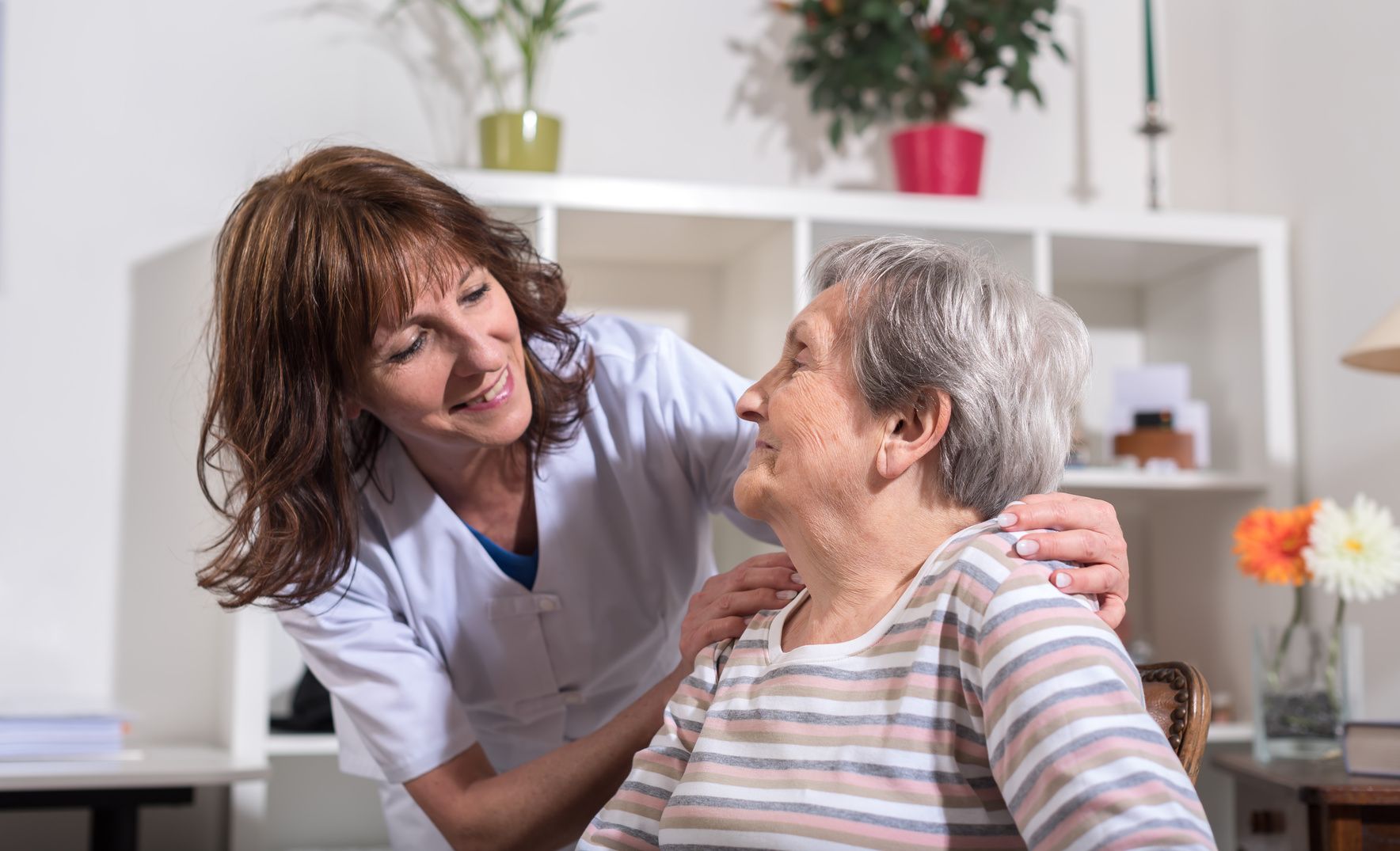 Une aide-soignante en uniforme blanc sourit et tient délicatement les épaules d'une femme âgée, toutes deux partageant un moment chaleureux à l'intérieur. La scène reflète des soins de qualité, avec des services à partir de 25 euros l'heure d'aide à domicile, conformément à l'arrêté du 23 décembre 2025 du SAD.