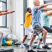 Un groupe de personnes âgées, hommes et femmes, s'exerce sur des plates-formes de marche dans un studio de gymnastique lumineux, levant les bras parallèlement au sol, tout en pratiquant ensemble une activité physique adaptée aux seniors.