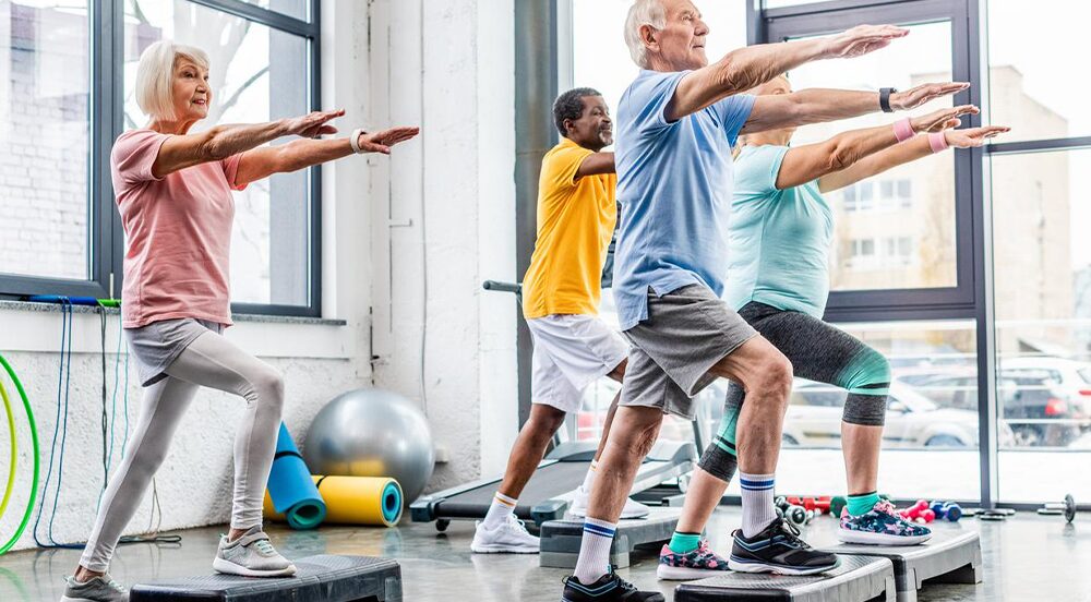 Un groupe de personnes âgées, hommes et femmes, s'exerce sur des plates-formes de marche dans un studio de gymnastique lumineux, levant les bras parallèlement au sol, tout en pratiquant ensemble une activité physique adaptée aux seniors.