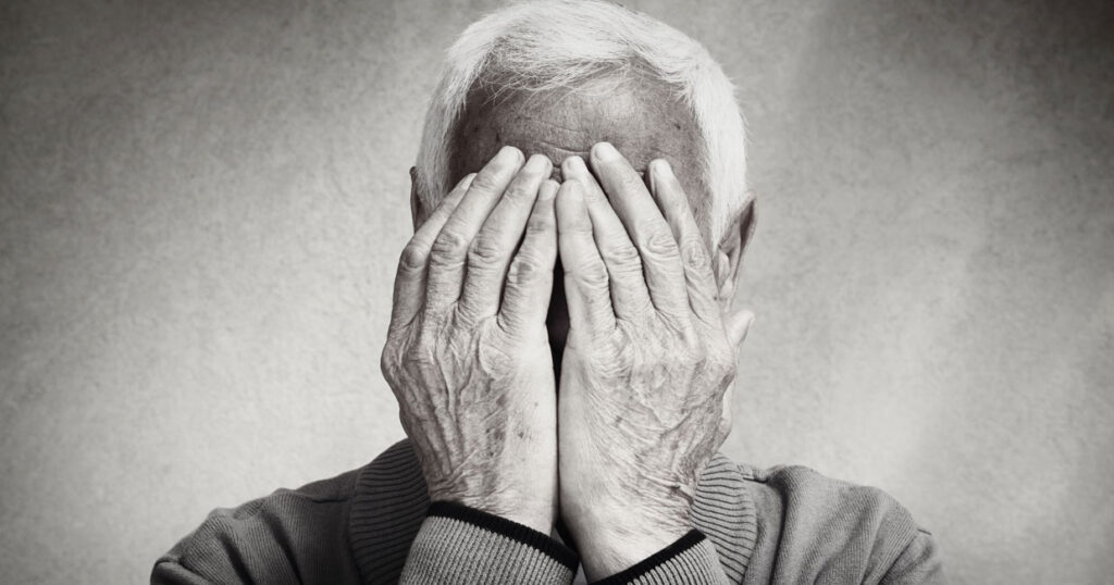 Une personne âgée aux cheveux blancs se couvre le visage des deux mains, ne laissant apparaître que ses mains et son front. Cette image en noir et blanc met en évidence les rides et la texture, capturant les signes dépression senior à travers une émotion subtile.
