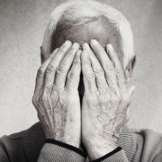 Une personne âgée aux cheveux blancs se couvre le visage des deux mains, ne laissant apparaître que ses mains et son front. Cette image en noir et blanc met en évidence les rides et la texture, capturant les signes dépression senior à travers une émotion subtile.