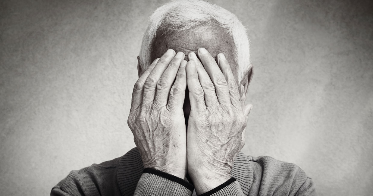 Une personne âgée aux cheveux blancs se couvre le visage des deux mains, ne laissant apparaître que ses mains et son front. Cette image en noir et blanc met en évidence les rides et la texture, capturant les signes dépression senior à travers une émotion subtile.