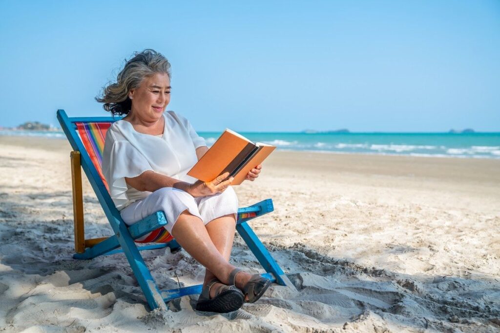 Une femme âgée est assise sur une chaise de plage colorée et lit un livre au bord de la mer pendant son séjour ANCV seniors en vacances, avec du sable blanc et un océan bleu en arrière-plan sous un ciel clair.