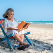 Une femme âgée est assise sur une chaise de plage colorée et lit un livre au bord de la mer pendant son séjour ANCV seniors en vacances, avec du sable blanc et un océan bleu en arrière-plan sous un ciel clair.